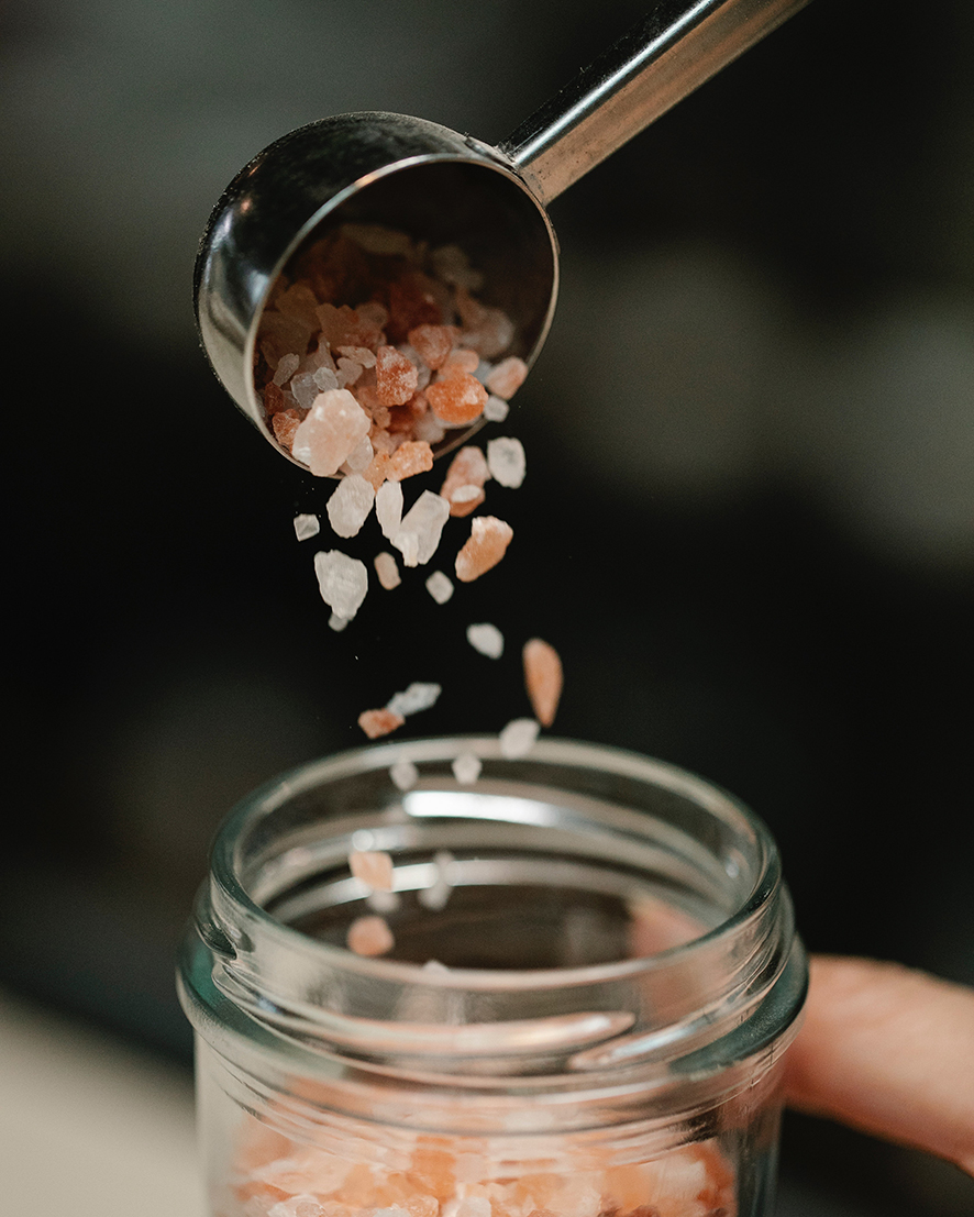 Himalájsku soľ použitú na výrobu domáceho mydla sype žena z kovovej odmerky do skleneného zatváracieho pohára.